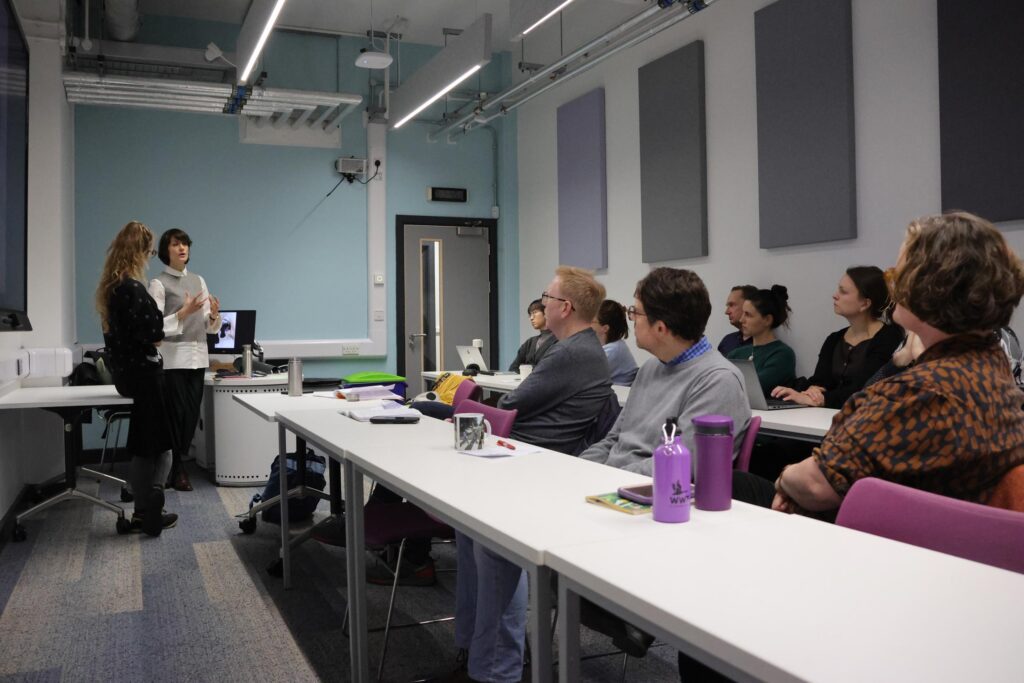 A small classroom setting where Dr Camille Bellet and Dr Eva Giraud stand at the front of the room addressing a group of adults seated at tables. The audience listens attentively, some with laptops and notebooks, in a modern room with light blue walls, overhead lighting, and acoustic panels.