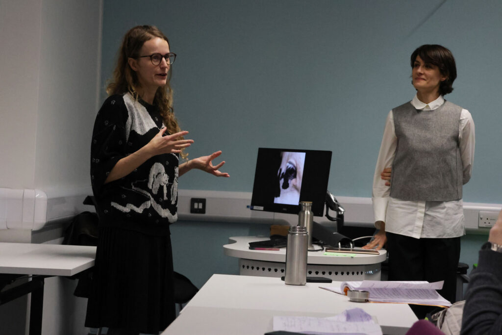 Dr Camille Bellet and Dr Eva Giraud stand at the front of a classroom: Eva gestures while introducing Camille, who stands beside her smiling. A computer monitor on the desk displays a photograph of a cow