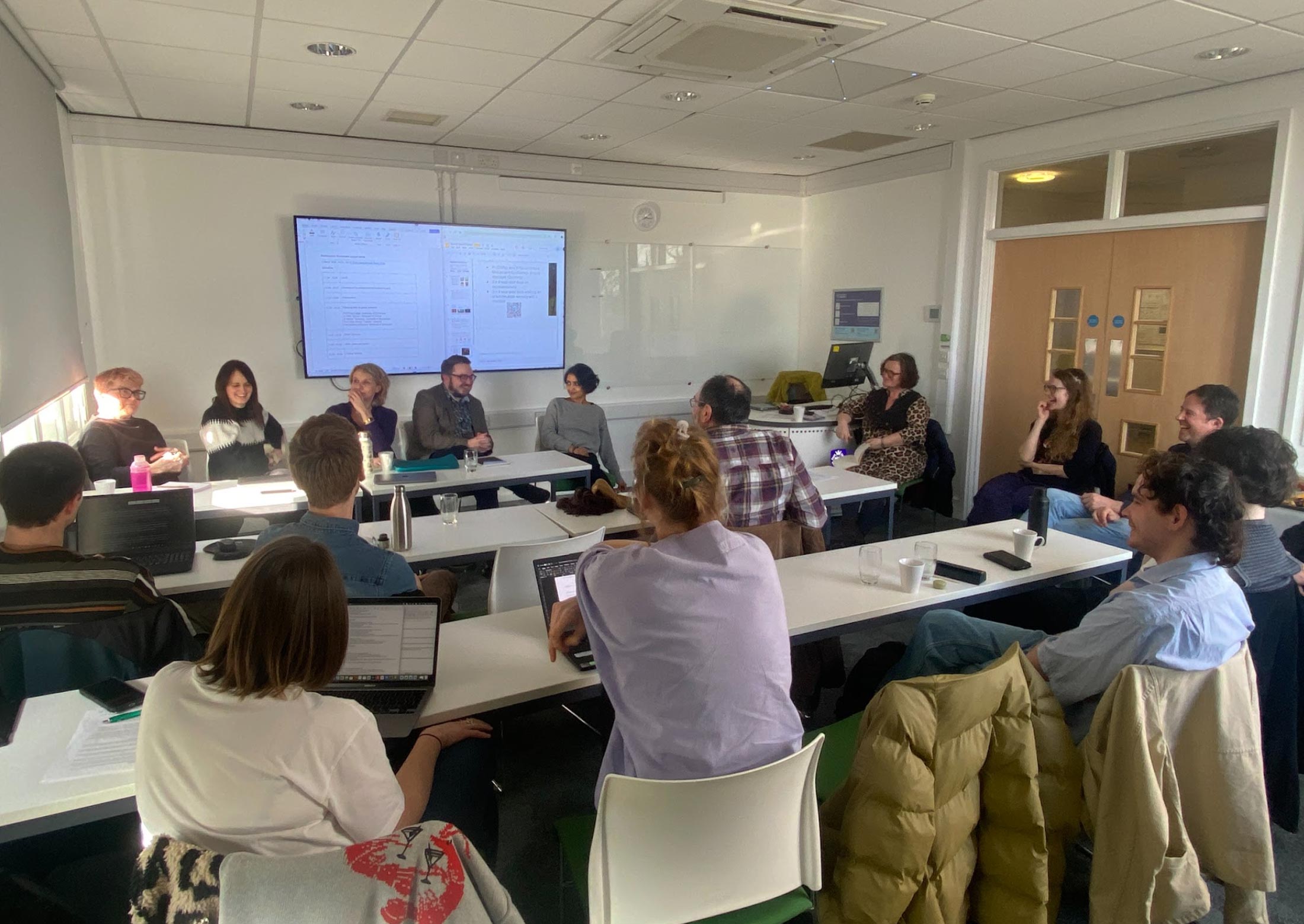 A group of adults sit in a bright meeting room, listening to the panelists at the front speak. A large screen behind the panel displays a document, and attendees have laptops, notebooks, and drinks on the tables.