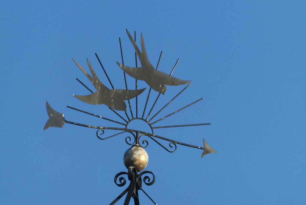 Iron weather vane mounted outdoors, silhouetted against a clear blue sky, indicating wind direction.