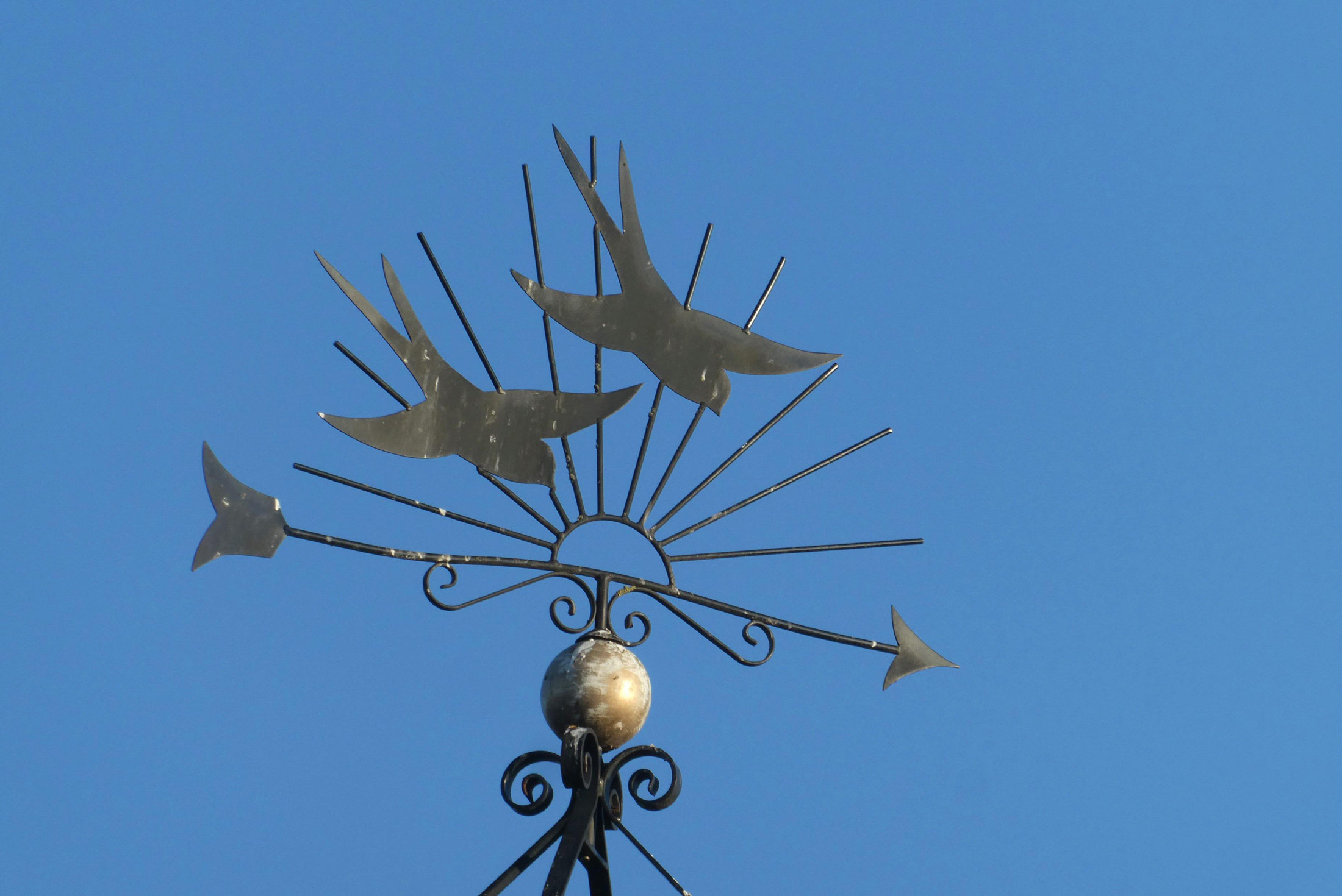 Iron weather vane mounted outdoors, silhouetted against a clear blue sky, indicating wind direction.