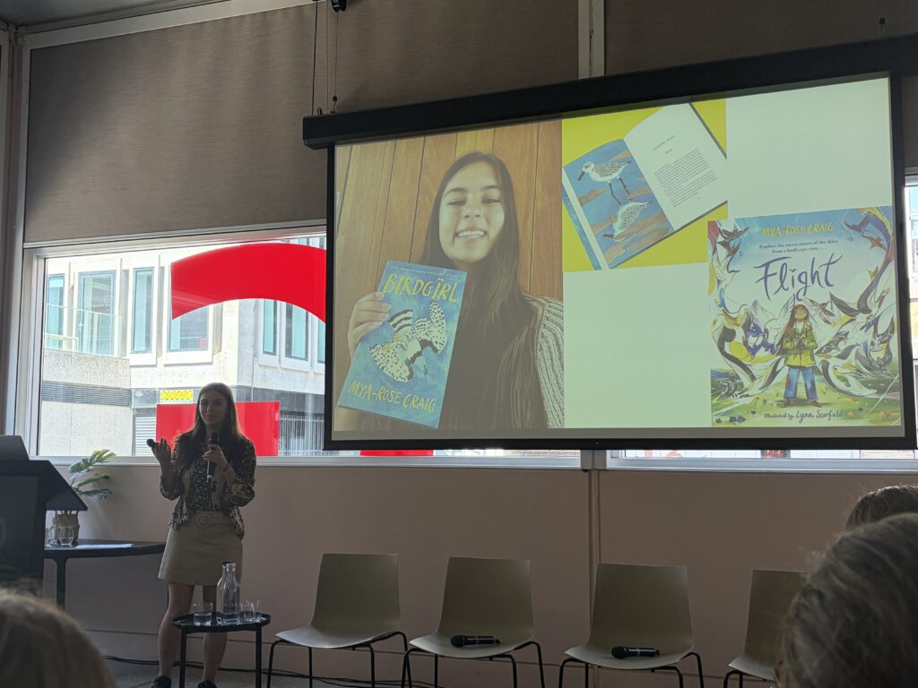 Dr Mya-Rose Craig stands at the front of a room speaking into a microphone. Behind her, a large projector screen displays slides featuring her books: Birdgirl and Flight, alongside a photo of her.