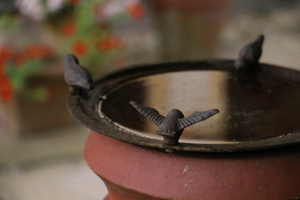 A close-up of a shallow metal bird bath mounted on a terracotta pedestal. Three small bird ornaments sit around the rim, with one in the foreground holding its wings open. The background is softly blurred with hints of flowers and greenery.