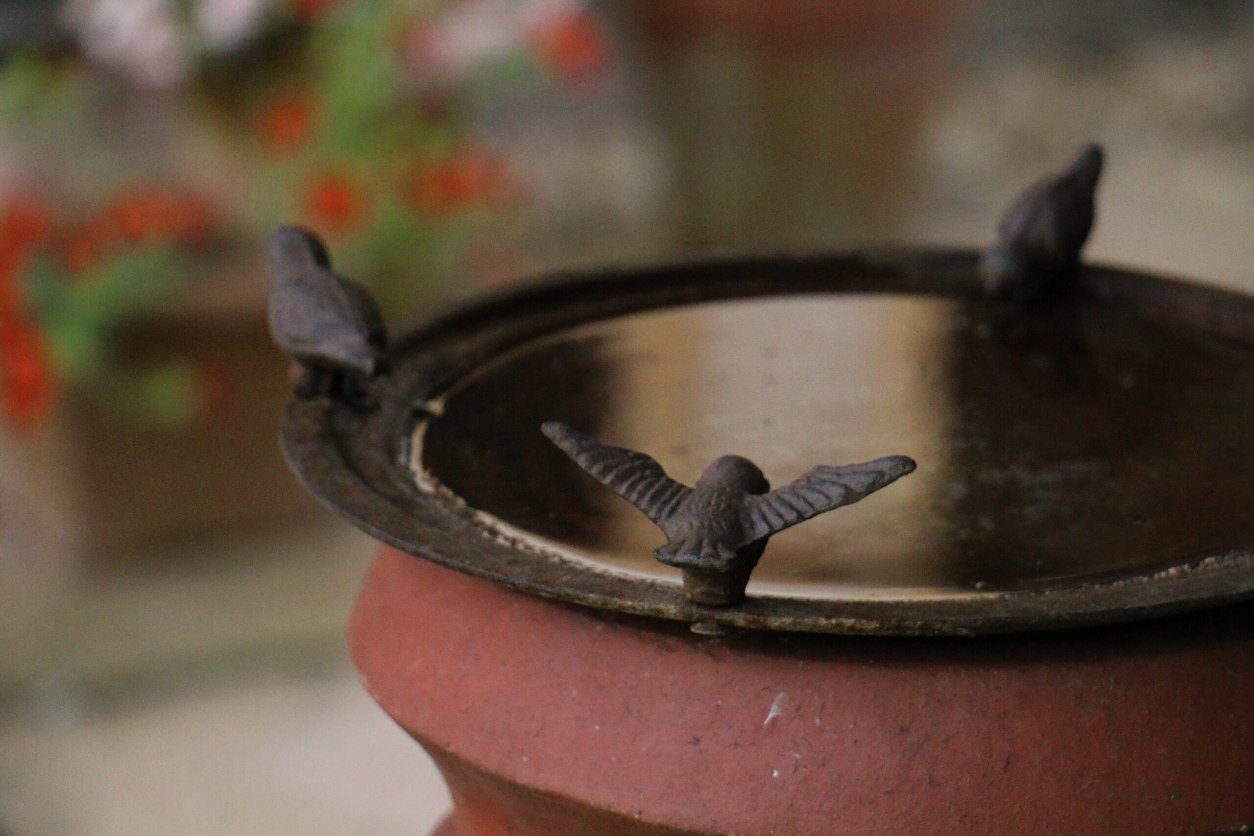 A close-up of a shallow metal bird bath mounted on a terracotta pedestal. Three small bird ornaments sit around the rim, with one in the foreground holding its wings open. The background is softly blurred with hints of flowers and greenery.
