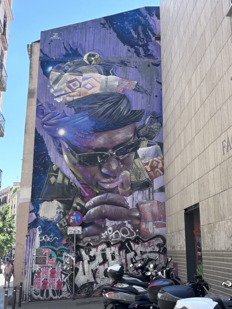 A vertical, multi-story mural painted on the side of a building in Barcelona. The mural features a large portrait of a person wearing black sunglasses, looking downward whilst giving a thumbs up sign. The figure is adorned with colorful patterns and a small, realistically detailed bird is perched on their shoulder.