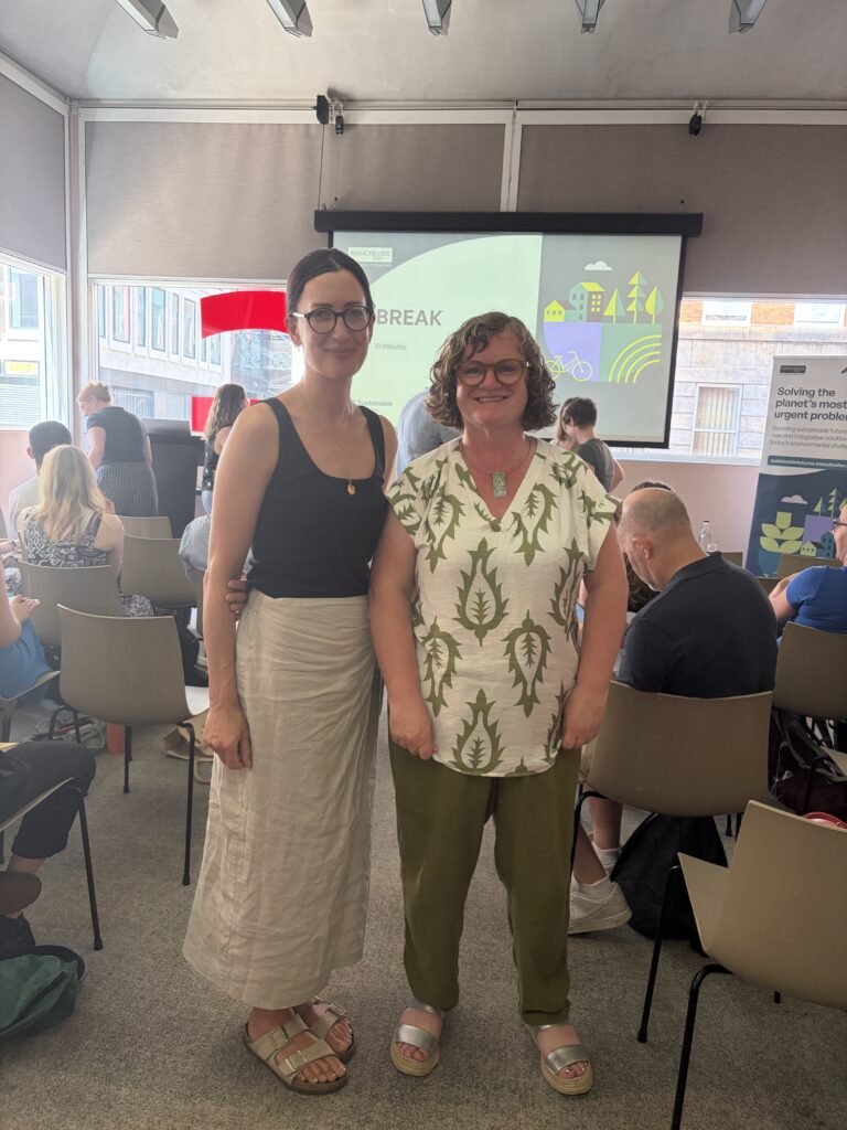 Lucy Dunning (left) and Rosaleen Duffy (right) stand smiling together in a conference room during a networking break at the Albatross lecture. Lucy wears a black sleeveless top and a light beige skirt and sandals, while Rosaleen wears a white blouse with a green leaf pattern, olive green trousers, and silver sandals. Behind them, attendees are seated and chatting, with a presentation screen displaying "BREAK" and sustainability-themed graphics, alongside a banner promoting sustainable futures in Manchester. Large windows on either side let in natural daylight.