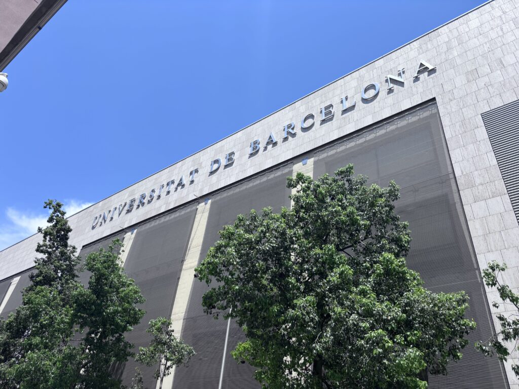 A photo of a building displaying the words ‘Universitat de Barcelona’ in silver metal signage against a bright blue sky. Large trees line the foreground.