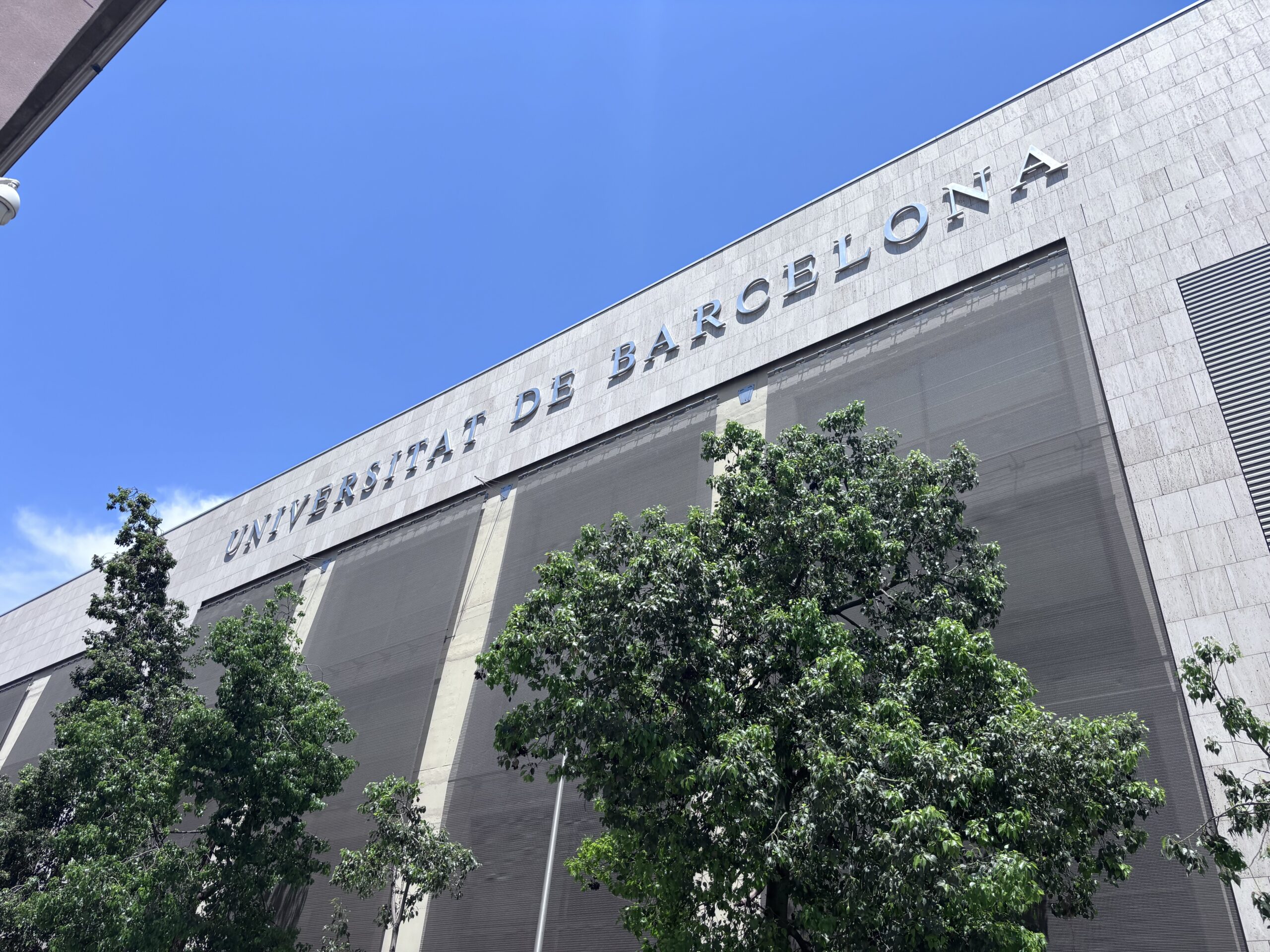 A photo of a building displaying the words ‘Universitat de Barcelona’ in silver metal signage against a bright blue sky. Large trees line the foreground.
