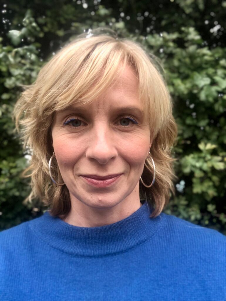 Maisie Tomlinson has blonde short hair and wears a blue jumper, pictured in front of a leafy background