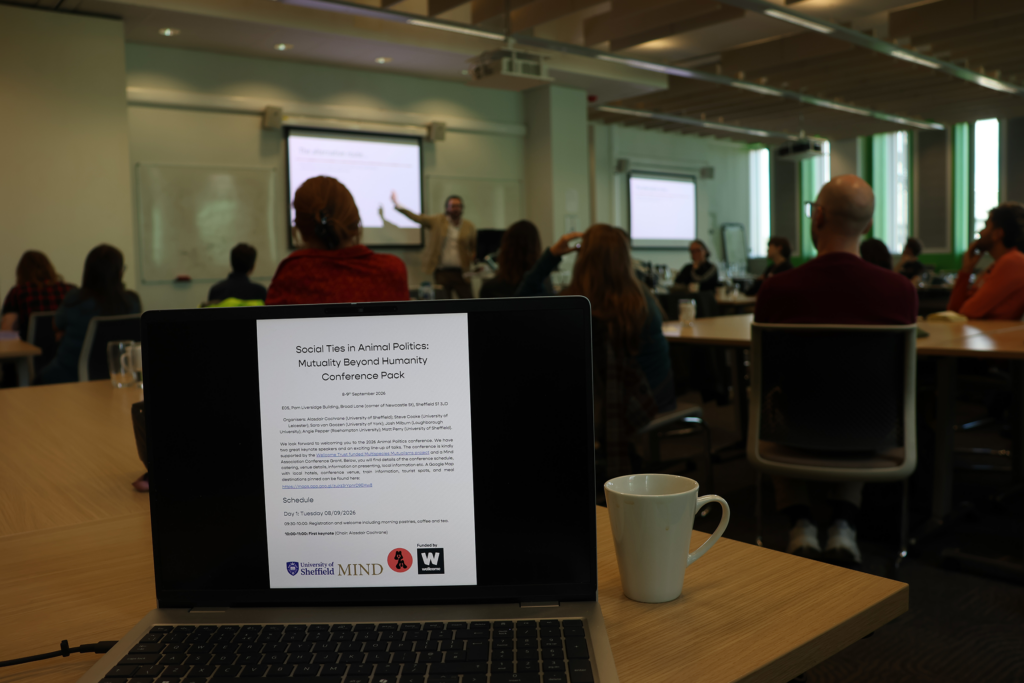 A laptop screen showing the conference pack in the foreground, with a presenter speaking to the audience in the background