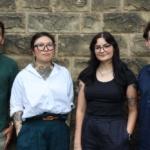 Four people standing side-by-side in a line against a stone wall, posing for a portrait.
