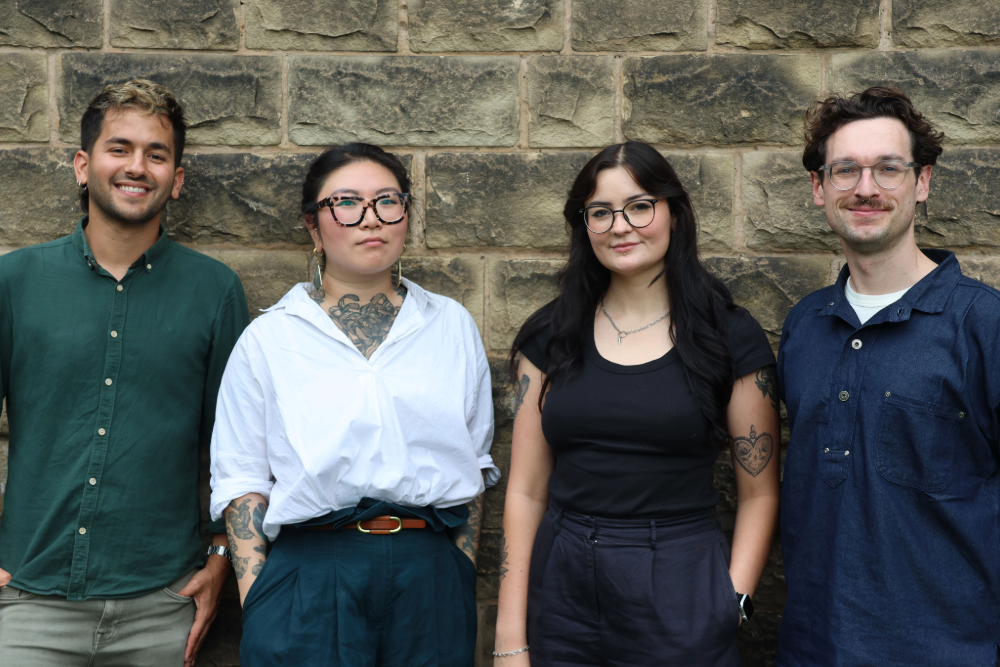 Four people standing side-by-side in a line against a stone wall, posing for a portrait.