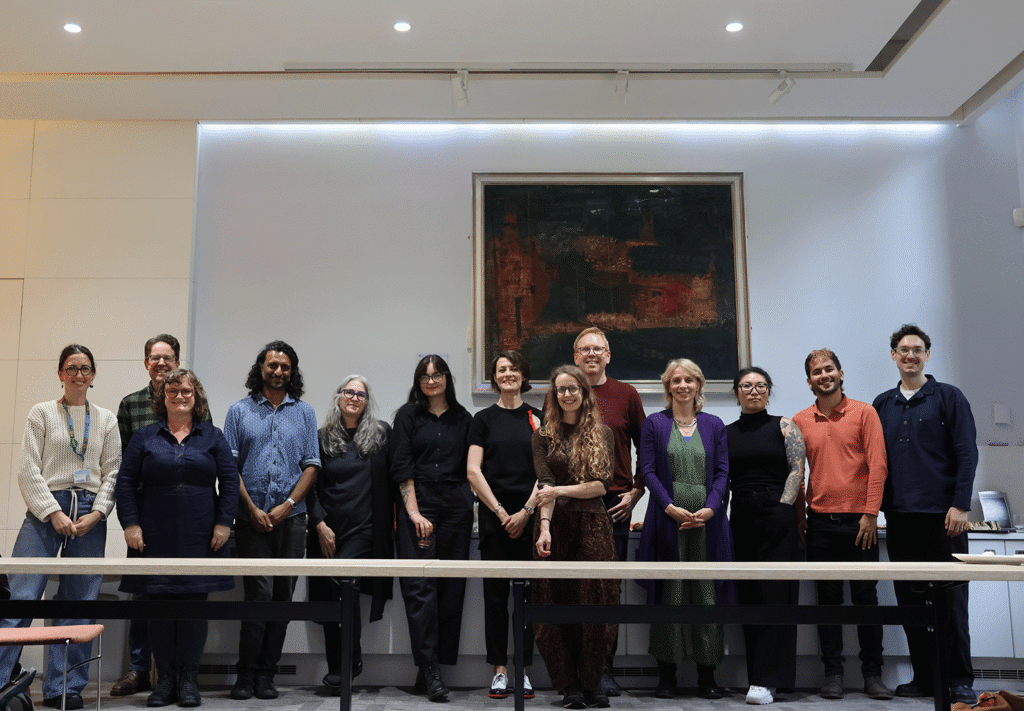A group photo of the team standing side-by-side in a line in a brightly lit modern room, with a large, framed dark oil painting hanging on the white wall behind them.