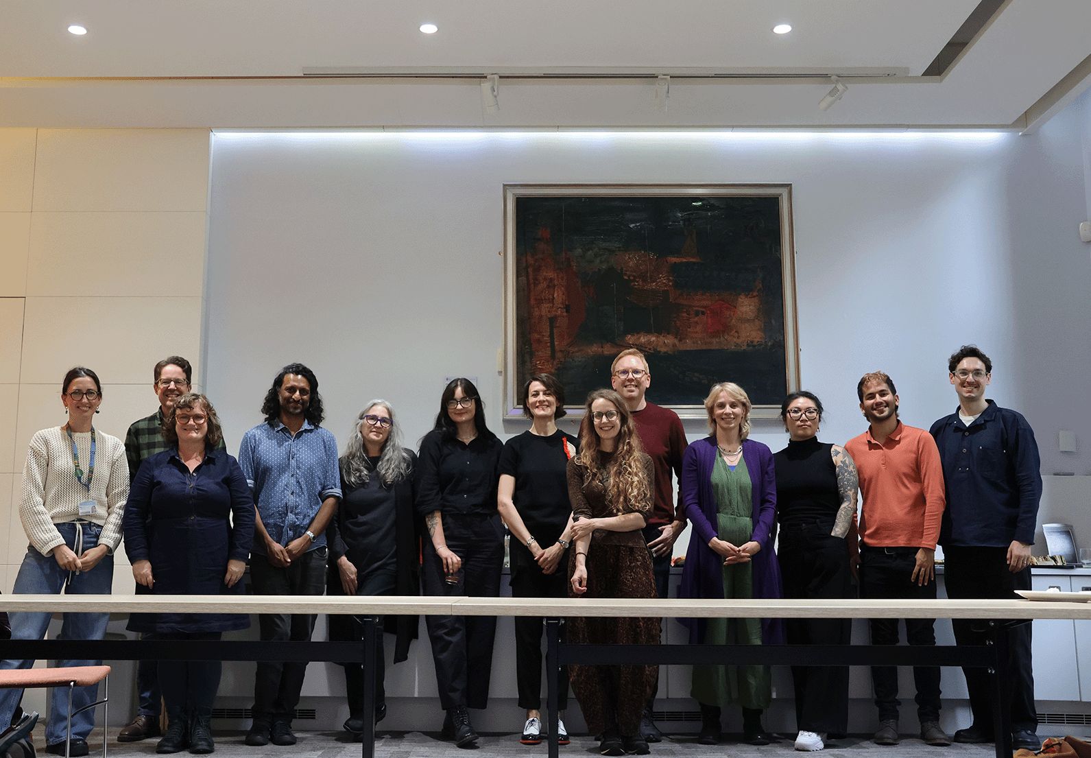 A group photo of the team standing side-by-side in a line in a brightly lit modern room, with a large, framed dark oil painting hanging on the white wall behind them.