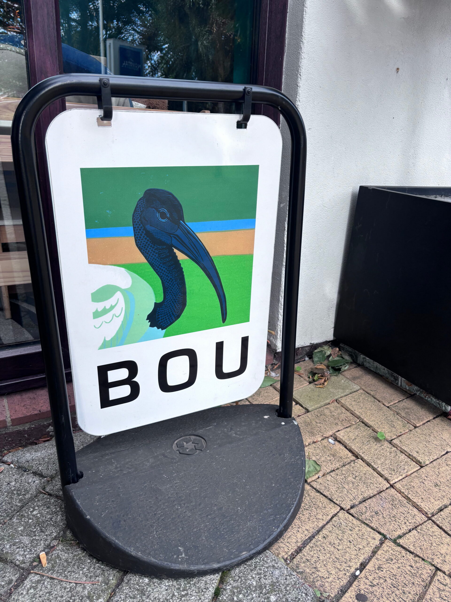 An outdoor A-frame pavement sign displaying a close-up graphic illustration of an ibis head against green and blue blocks with the letters "BOU" underneath.